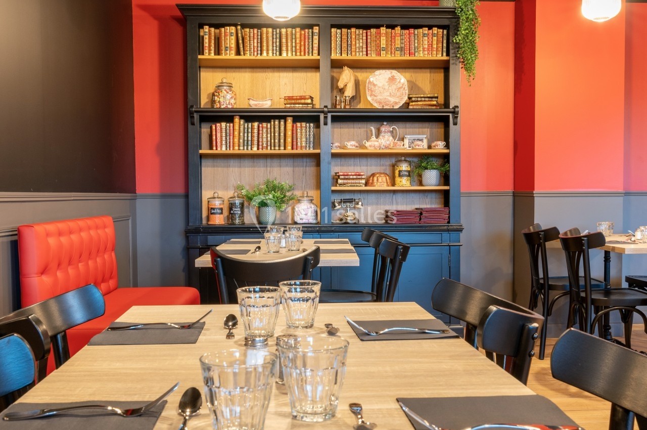 Salle de restaurant avec tables dressées, chaises noires, banquette rouge et étagère décorative en arrière-plan.