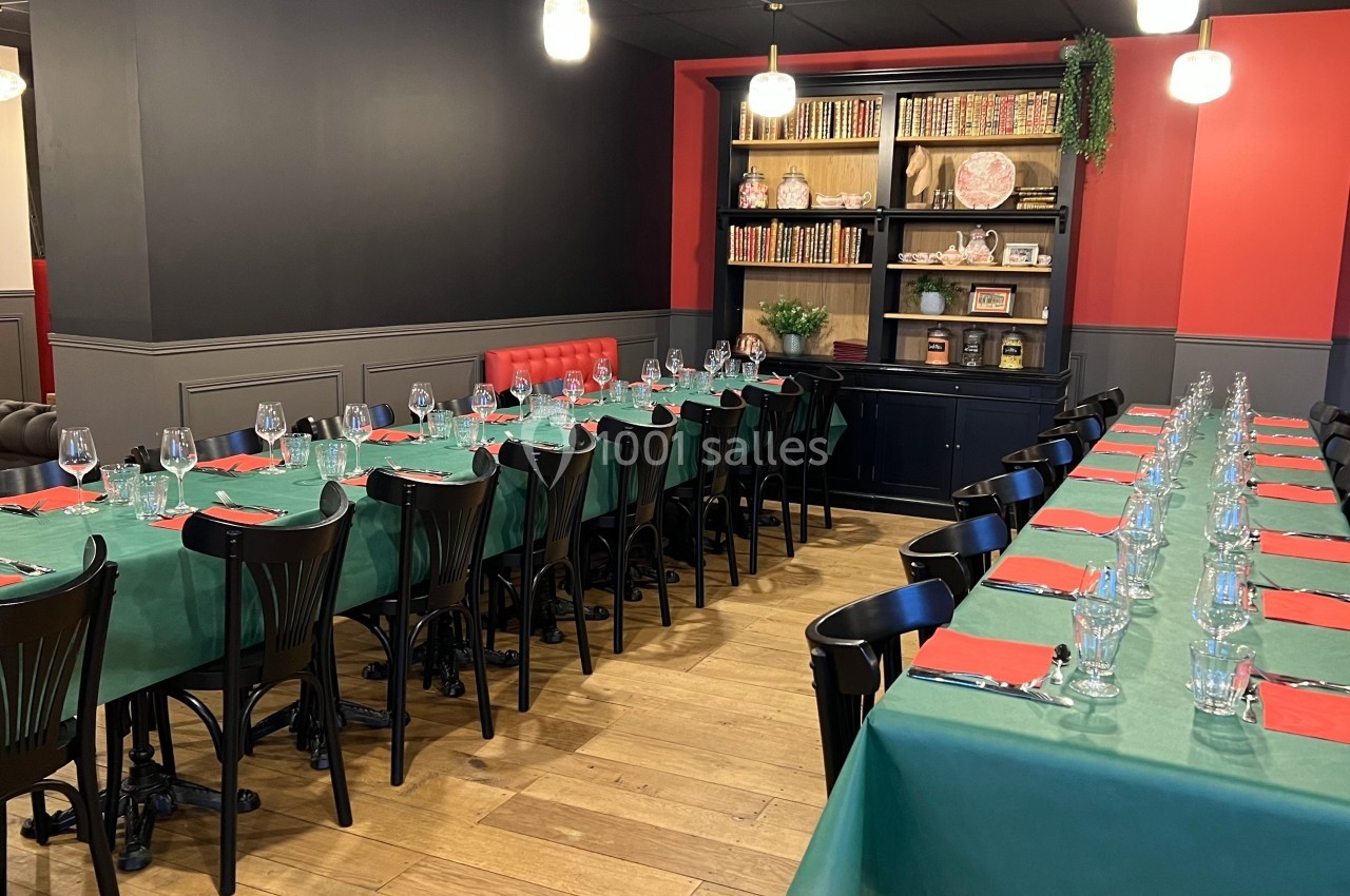 Salle de restaurant aménagée avec deux longues tables dressées, nappes vertes, serviettes rouges et étagères en arrière-plan.