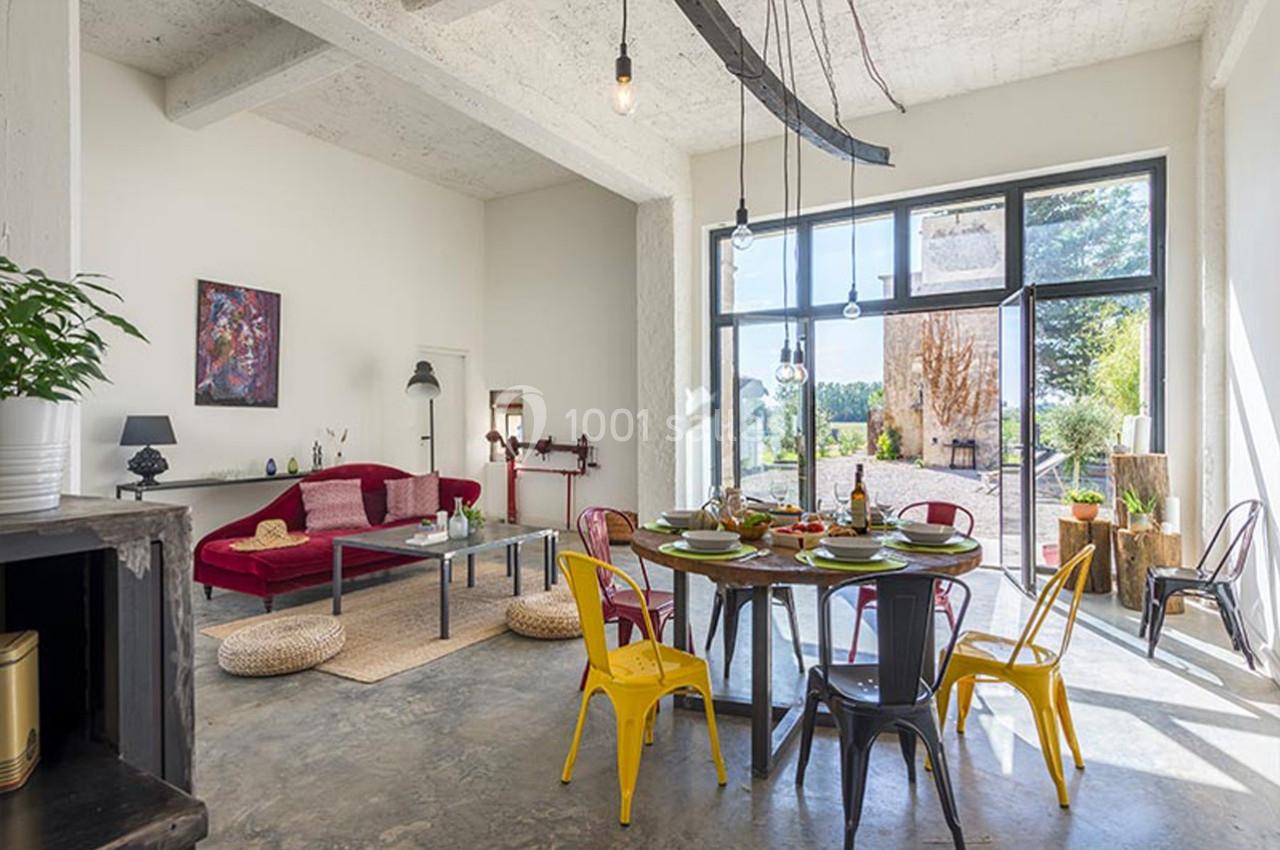 Salle à manger lumineuse avec table dressée, chaises colorées, canapé rouge et grandes baies vitrées donnant sur un jardin.