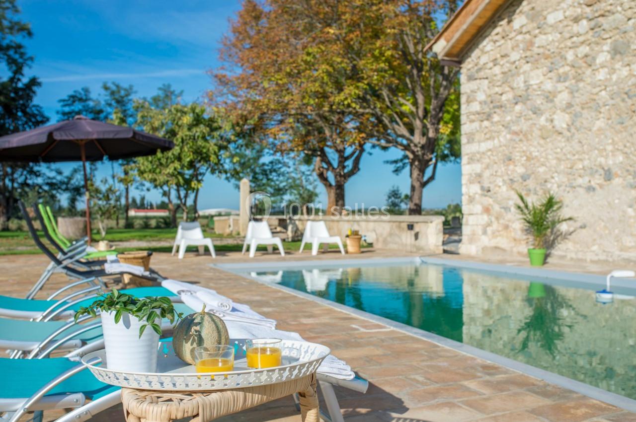 Terrasse avec piscine, transats, parasol et boissons sur une table, entourée d'arbres et d'un bâtiment en pierre.