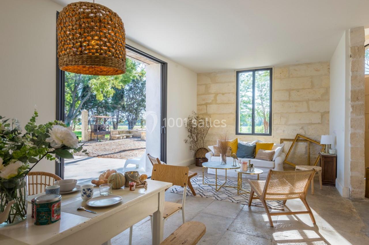 Salon lumineux avec murs en pierre, mobilier en bois clair, grandes fenêtres ouvertes sur un jardin arboré.