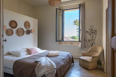 Chambre lumineuse avec lit double, fauteuil beige, fenêtre ouverte sur un paysage et décoration murale en osier.