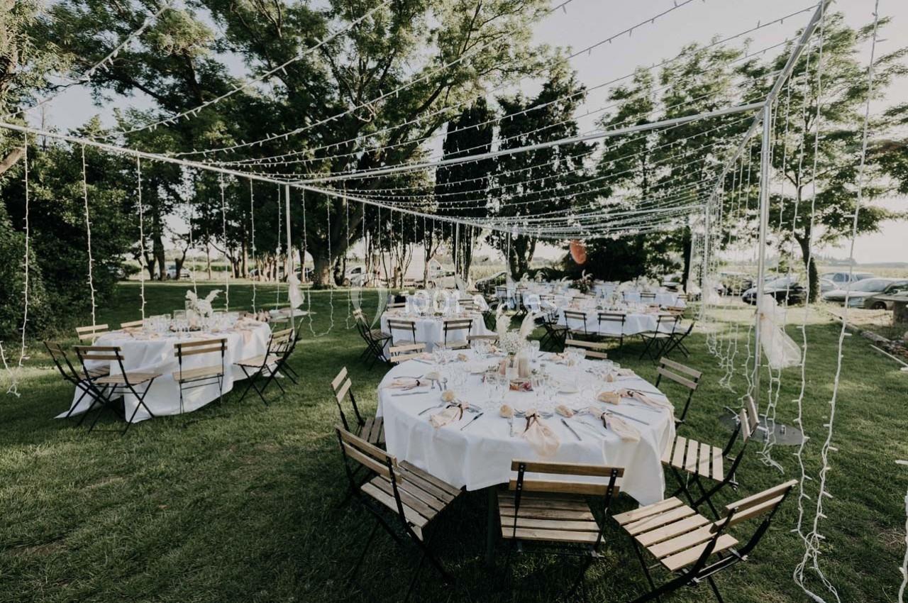 Tables rondes dressées avec nappes blanches et chaises pliantes en bois, disposées sous des guirlandes lumineuses dans un…