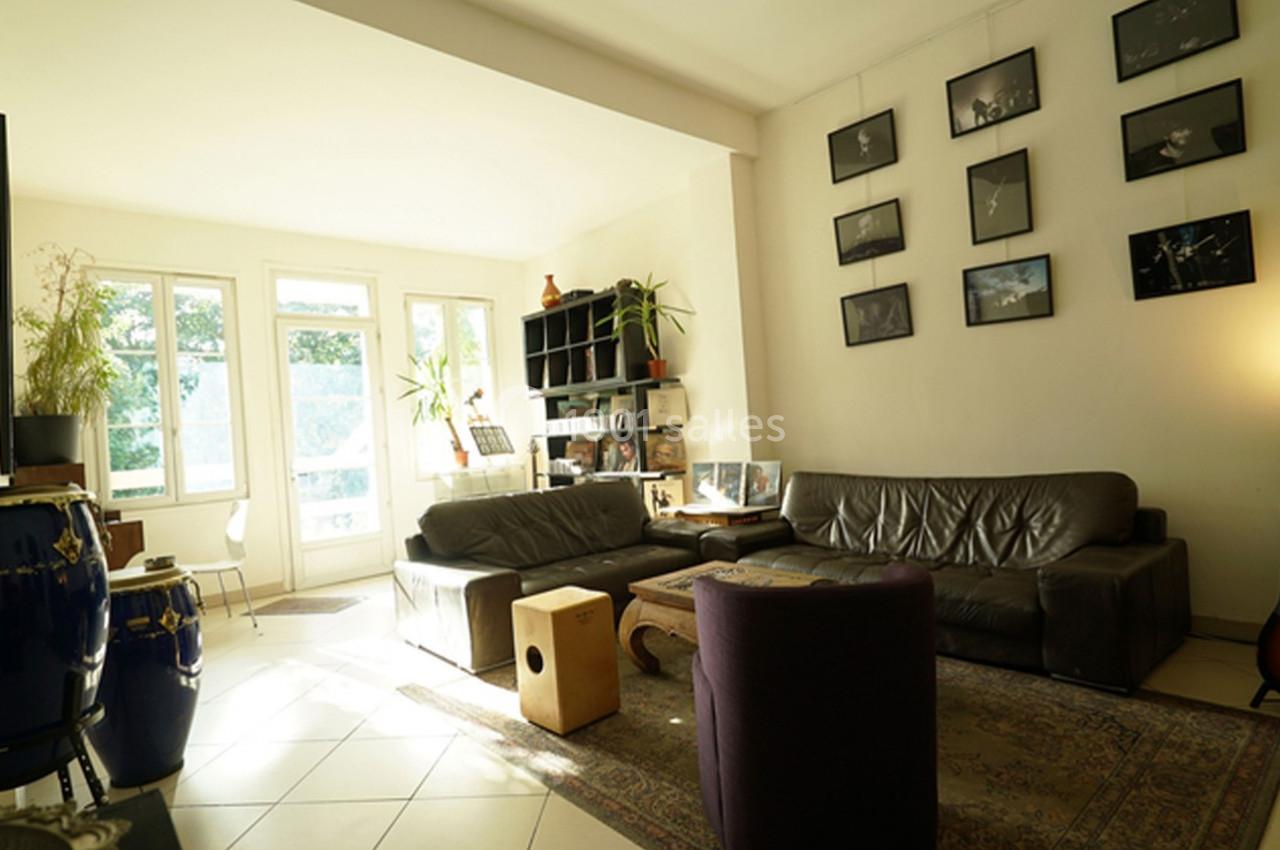 Salon lumineux avec canapé en cuir, instruments de musique, étagères, photos encadrées au mur et grandes fenêtres.