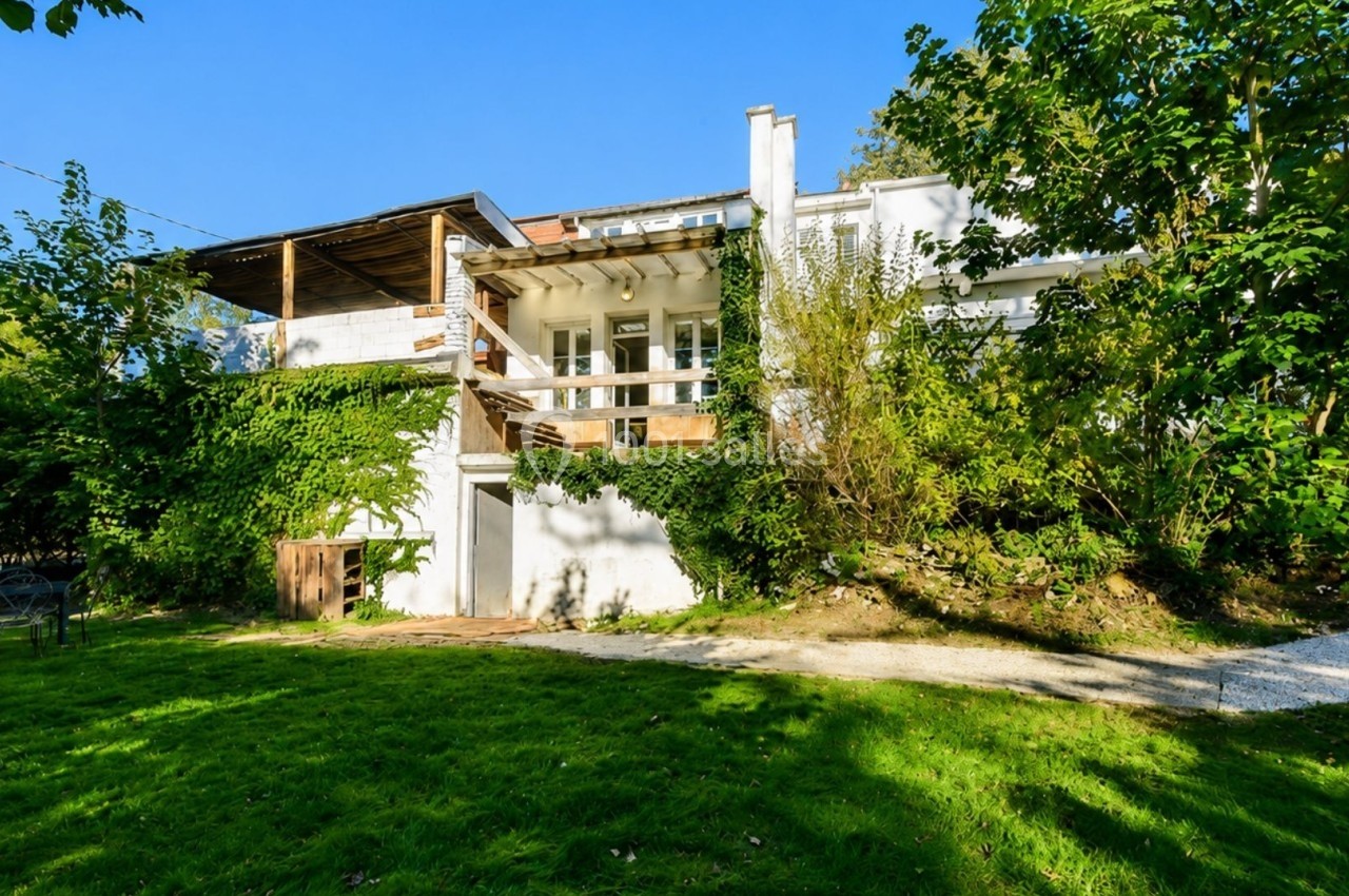Façade d'une maison blanche entourée de végétation, avec terrasse en bois et jardin verdoyant au premier plan.