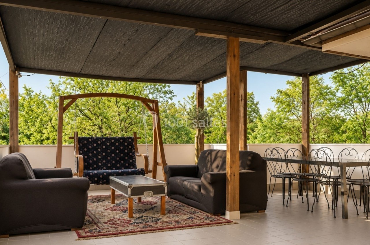 Terrasse couverte avec salon de jardin, balançoire en bois, table et chaises en métal, entourée de verdure.