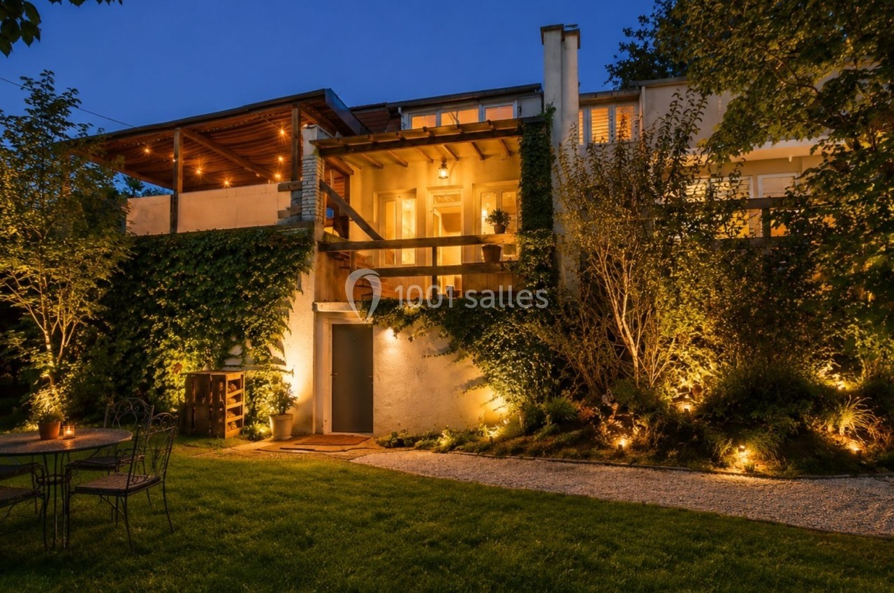 Maison illuminée en soirée avec terrasse en bois, jardin aménagé et mobilier extérieur sur pelouse.
