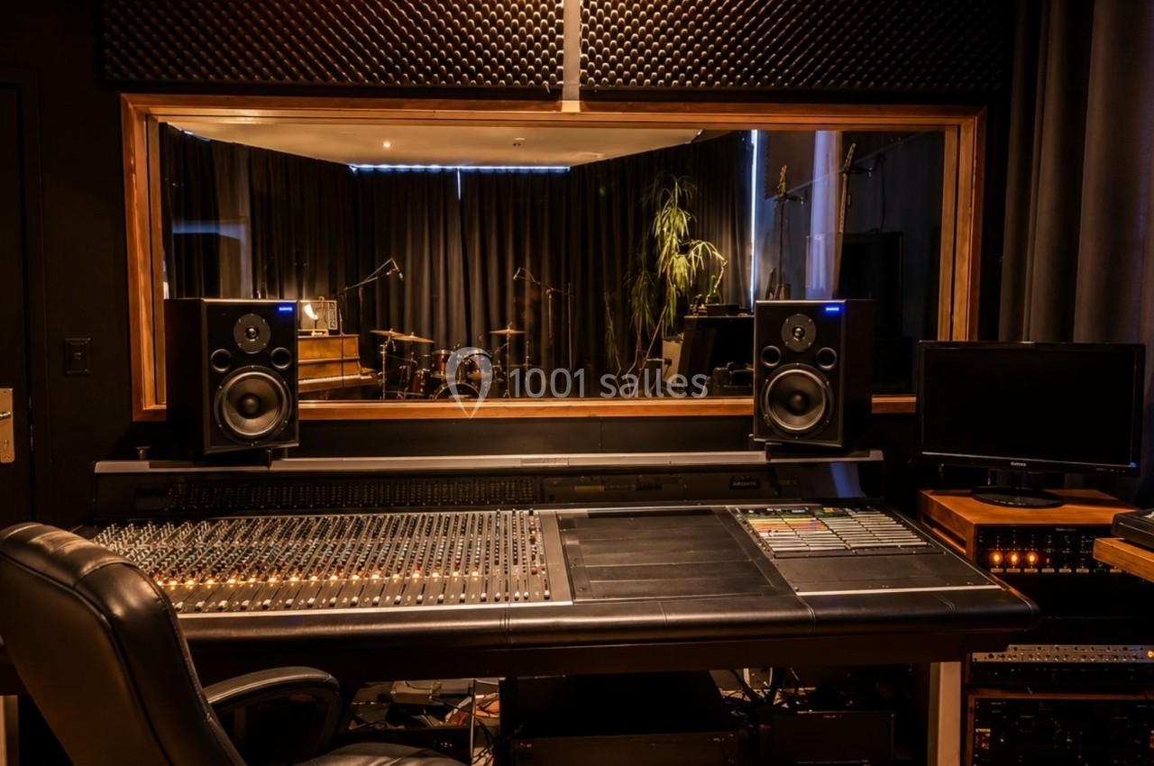 Salle de studio d'enregistrement avec table de mixage, enceintes, écran et vue sur une pièce avec instruments de musique.