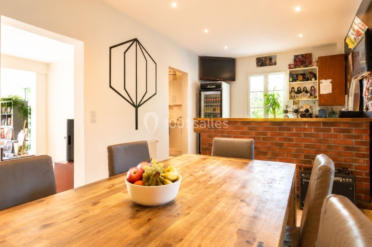 Salle à manger lumineuse avec table en bois, chaises en cuir, et cuisine ouverte avec bar en briques au fond.