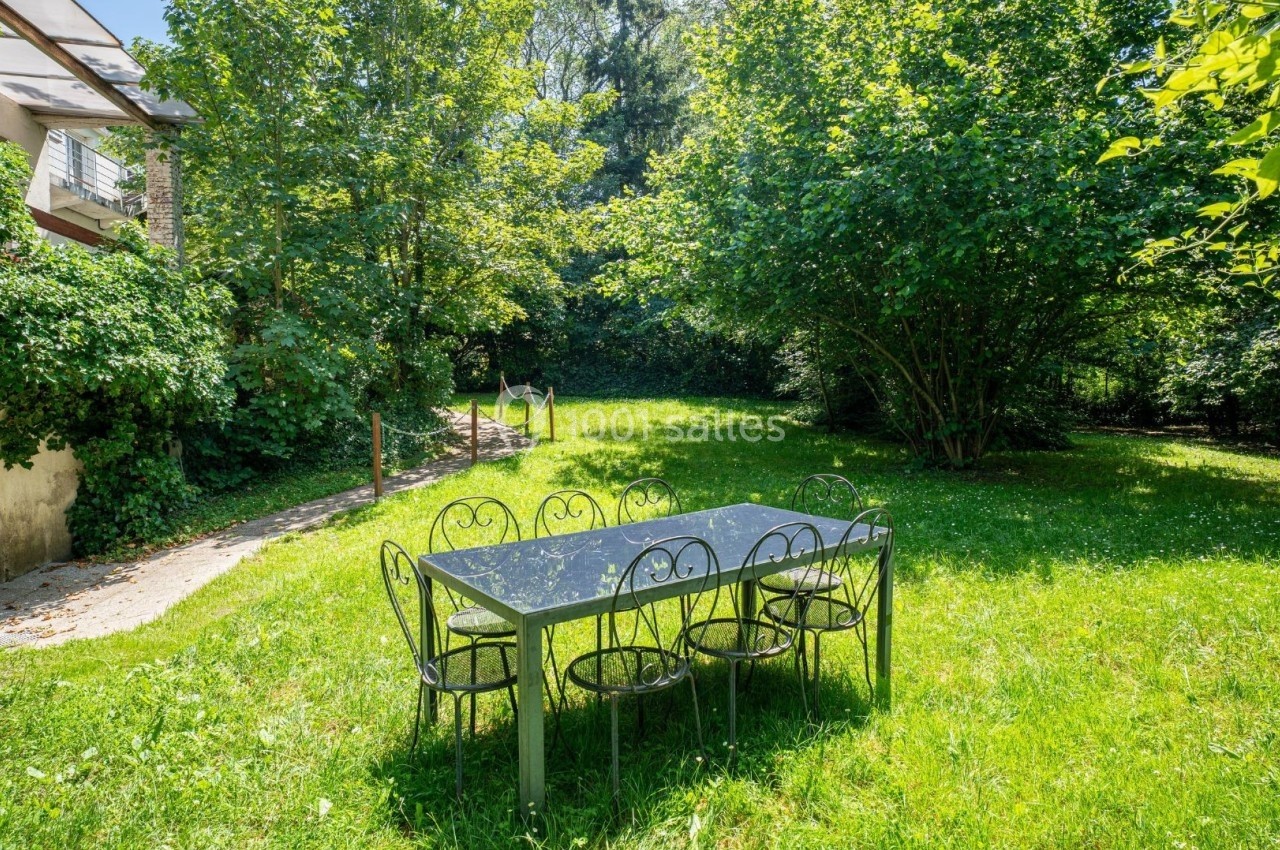 Table et chaises en métal noir disposées sur une pelouse verdoyante, entourées d'arbres et de végétation dense.