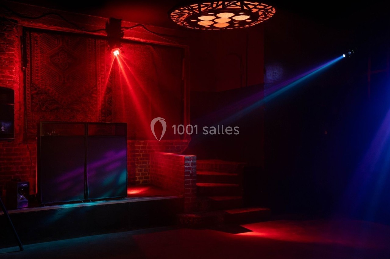 Salle sombre avec éclairage rouge et bleu, une estrade avec équipement audio et un tapis mural en arrière-plan.