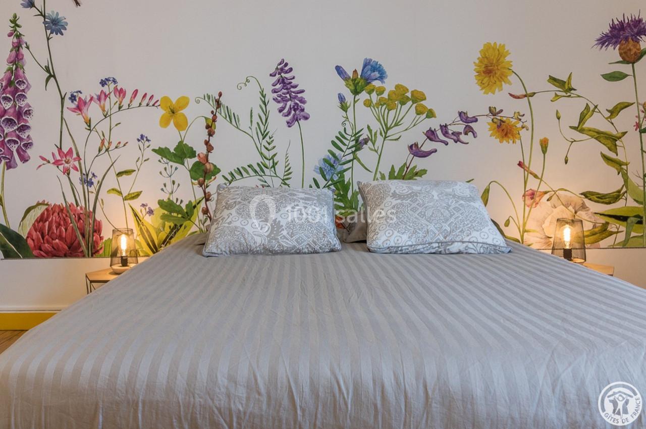 Lit double avec housse grise, encadré de tables de chevet et éclairé par des lampes, devant un mur décoré de fleurs colorées.