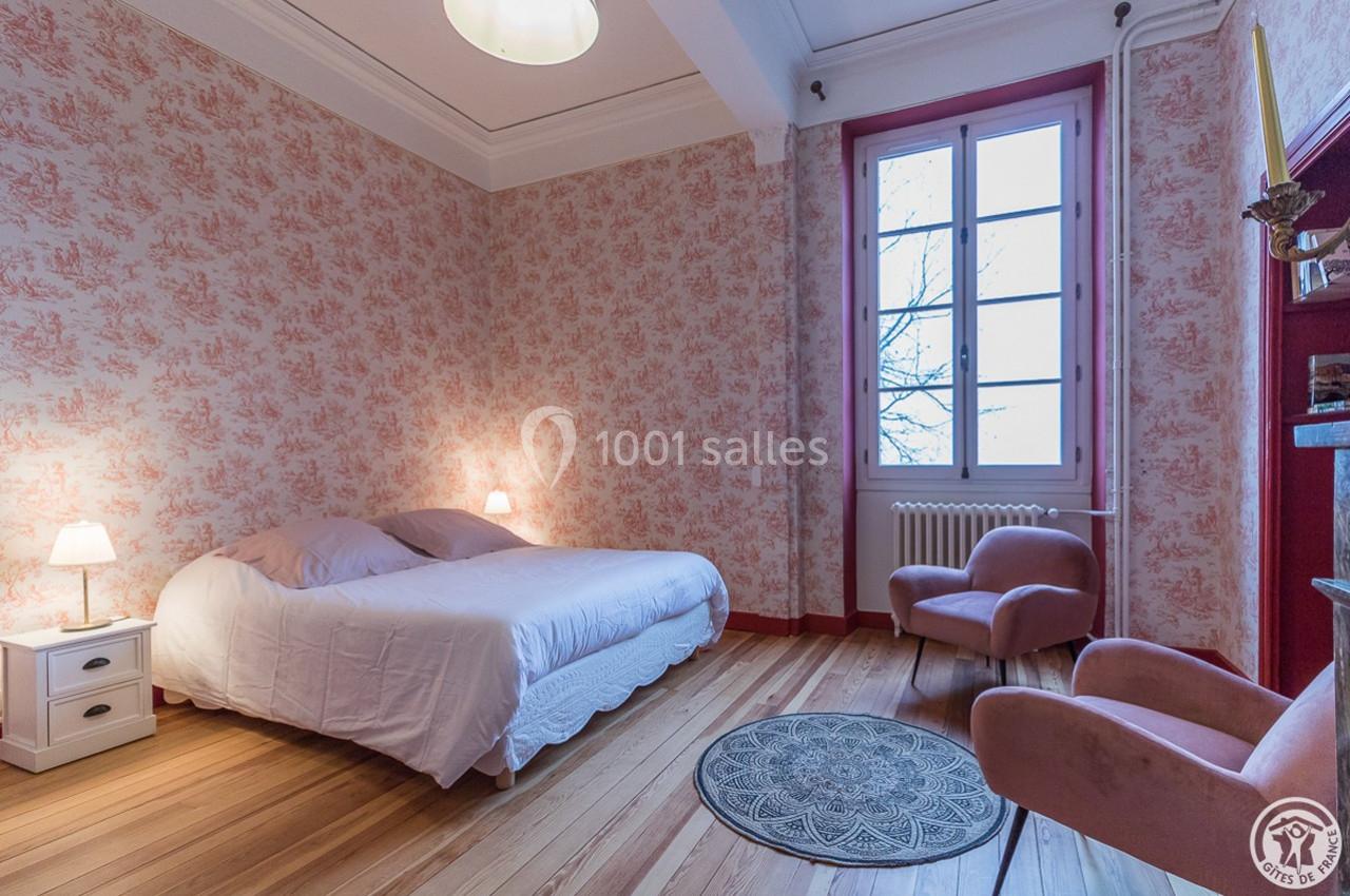 Chambre avec lit double, fauteuils roses, papier peint floral, parquet en bois et grande fenêtre laissant entrer la lumière.