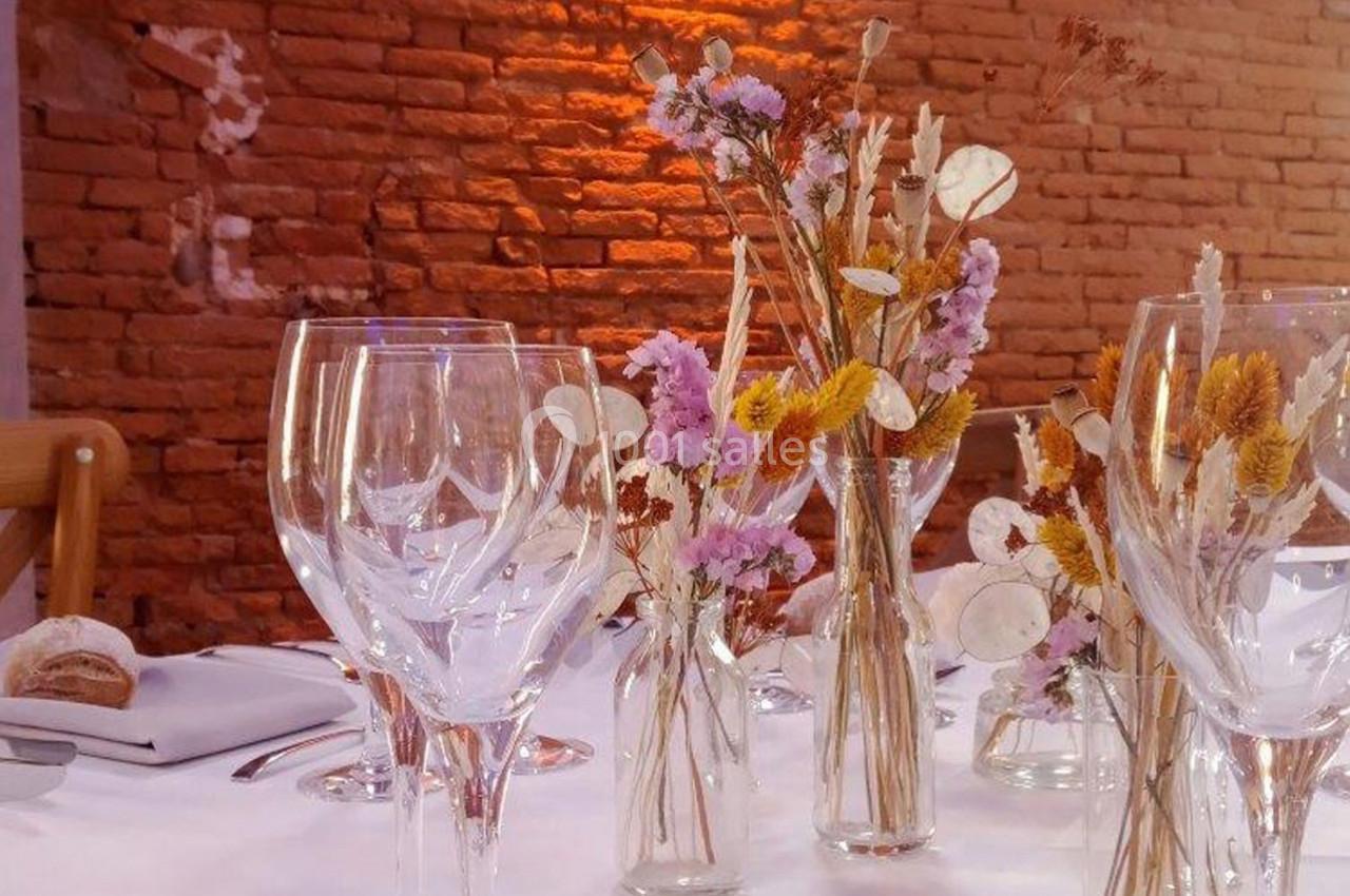 Table décorée avec des fleurs séchées dans des vases en verre, entourée de verres à vin, devant un mur en briques.