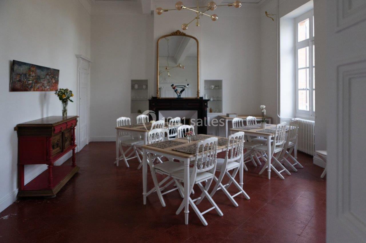 Salle lumineuse avec tables et chaises blanches, grand miroir doré, buffet rouge et sol en carrelage rouge.