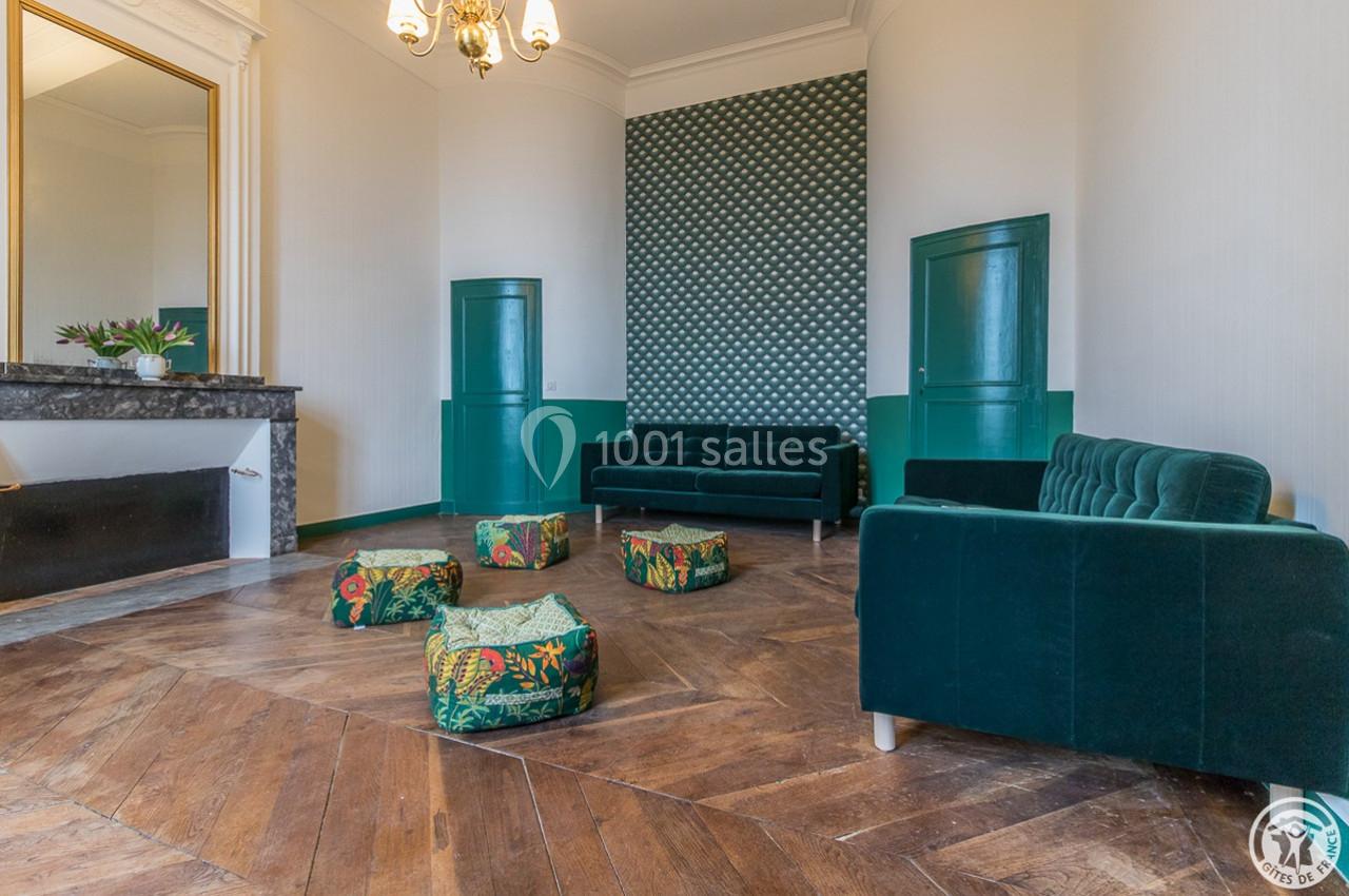 Salon lumineux avec parquet en bois, canapés verts, poufs à motifs tropicaux et cheminée décorative.