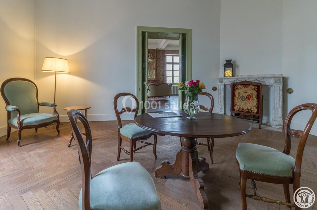 Salle à manger avec table ronde en bois, chaises anciennes, parquet, lampe, cheminée et bouquet de fleurs.