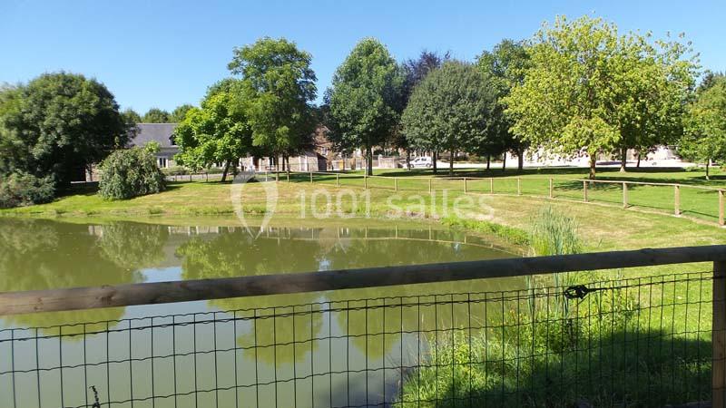 Étang entouré de pelouse, clôture en bois et arbres, avec des bâtiments en arrière-plan sous un ciel dégagé.