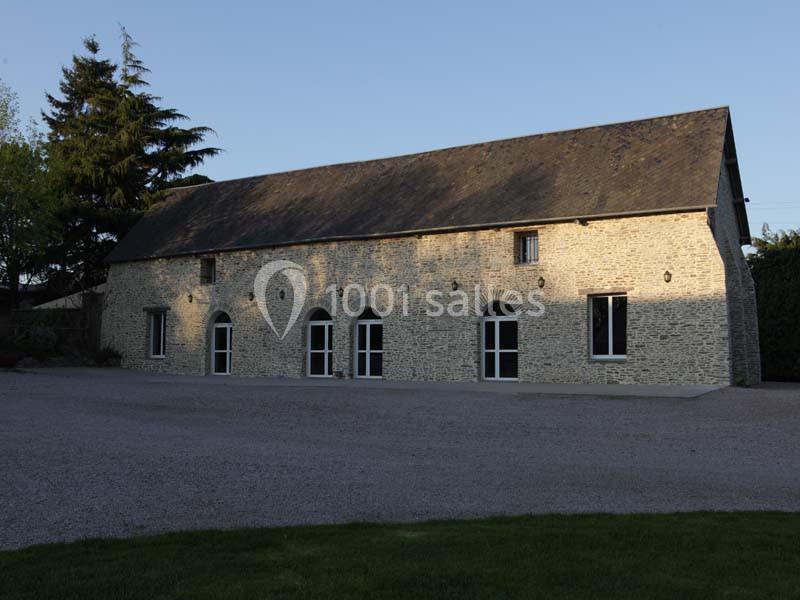 Bâtiment en pierre avec toit en ardoise, grandes portes vitrées et arbres en arrière-plan, sous un ciel dégagé.