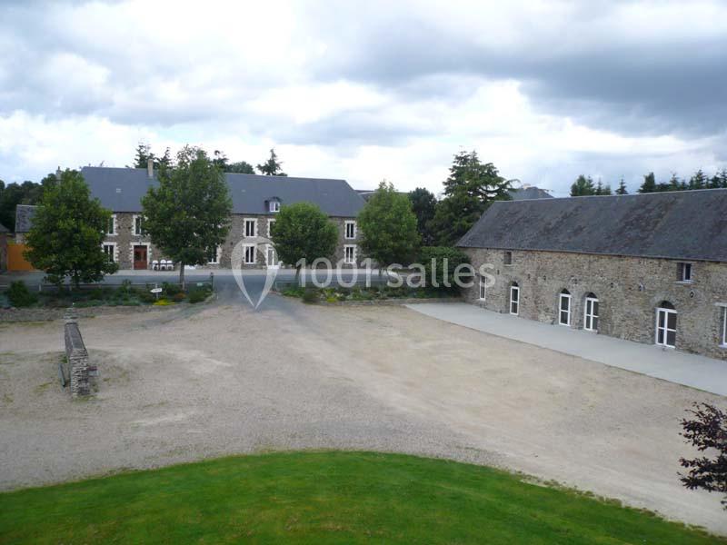 Cour d'un domaine avec bâtiments en pierre, arbres alignés et ciel nuageux en arrière-plan.