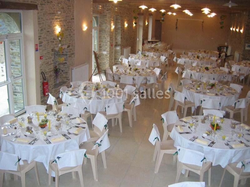 Salle de réception décorée avec des tables rondes dressées pour un événement, chaises blanches et murs en pierre apparente.