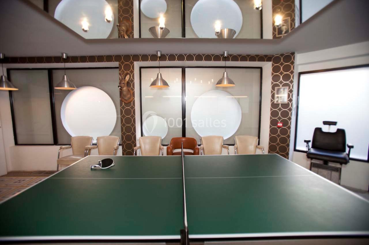 Salle de réunion moderne avec une table de ping-pong au centre, entourée de chaises et éclairée par des suspensions.
