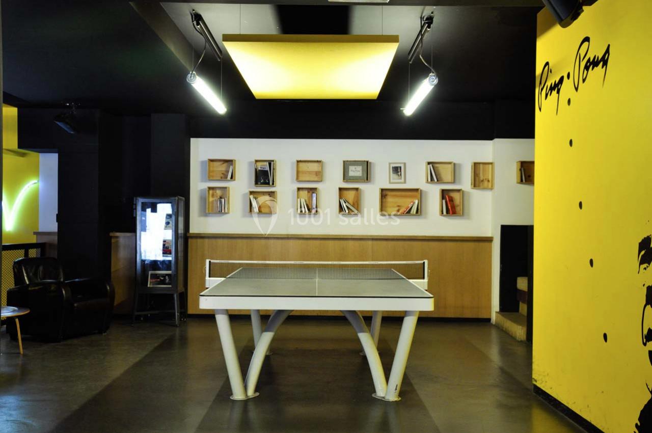 Table de ping-pong dans une pièce moderne avec étagères murales, fauteuil et décorations jaunes et noires.