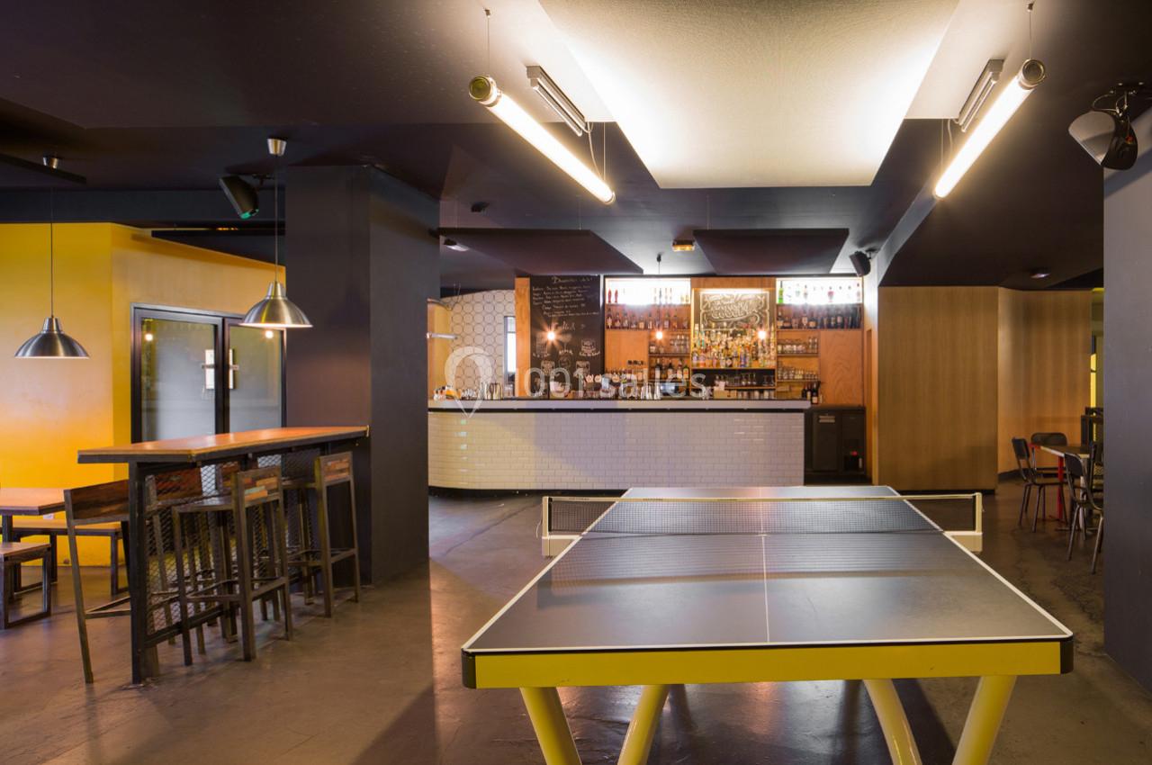 Salle moderne avec un bar éclairé, une table de ping-pong au premier plan et des tables hautes en arrière-plan.