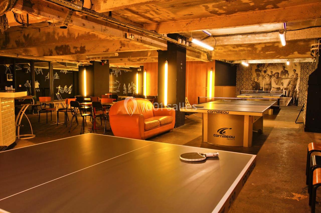 Salle de loisirs avec tables de ping-pong, canapé orange, chaises et éclairage chaleureux dans un espace industriel.