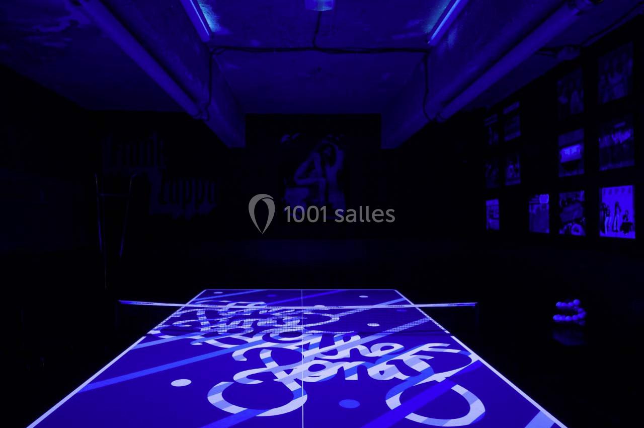 Table de ping-pong éclairée par une lumière noire, avec des motifs lumineux et des affiches en arrière-plan.