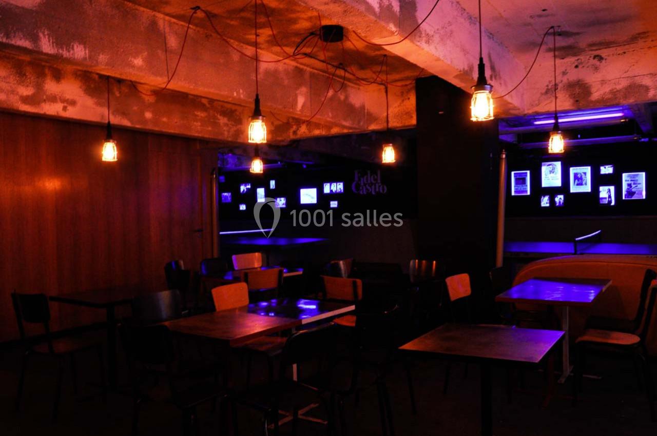 Salle sombre avec des tables et chaises vides, éclairée par des ampoules suspendues et des lumières violettes.