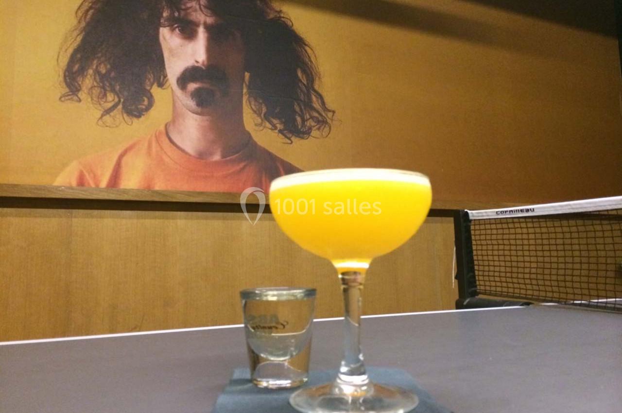 Un cocktail orange et un verre à shot posés sur une table de ping-pong, avec un portrait mural en arrière-plan.