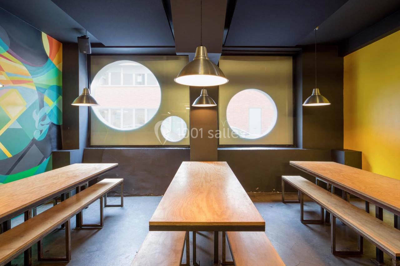 Salle moderne avec tables en bois, bancs assortis, murs colorés et grandes fenêtres rondes laissant entrer la lumière.