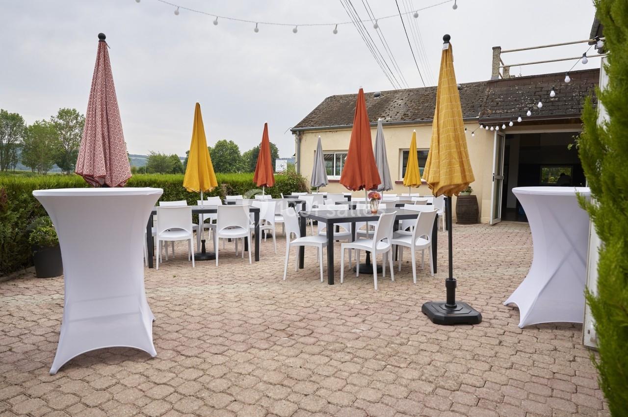 Terrasse extérieure avec tables, chaises blanches et parasols colorés, entourée de verdure et éclairée par des guirlandes.