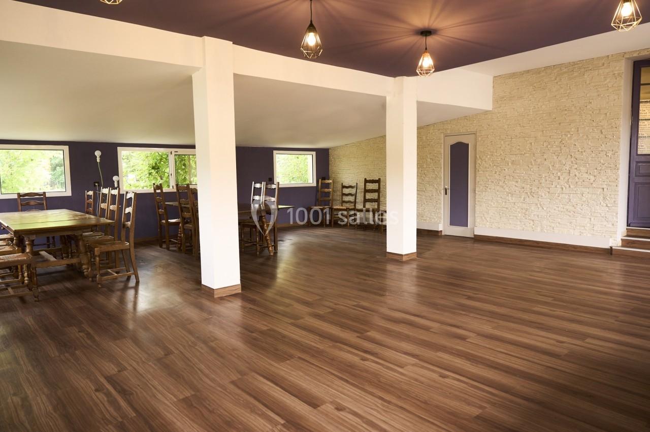 Salle spacieuse avec parquet en bois, murs blancs et violets, éclairage suspendu et grandes fenêtres donnant sur la verdure.