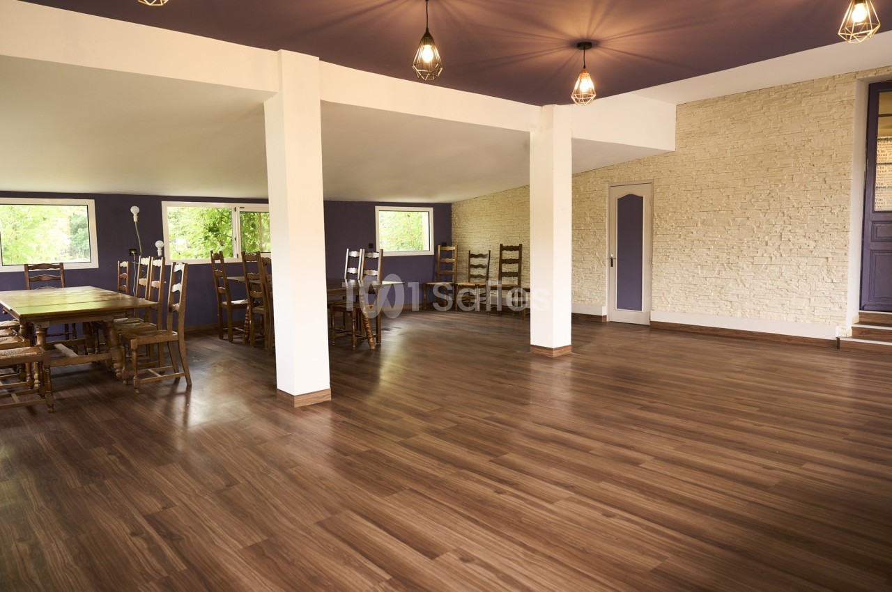 Salle spacieuse avec parquet en bois, murs blancs et violets, éclairage suspendu et grandes fenêtres donnant sur la verdure.