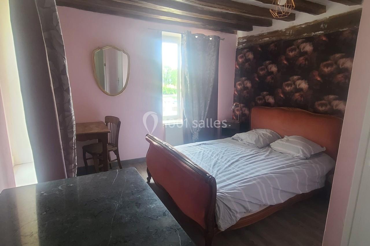 Chambre avec lit double, murs roses, poutres apparentes, table en bois, miroir ovale et fenêtre donnant sur l'extérieur.