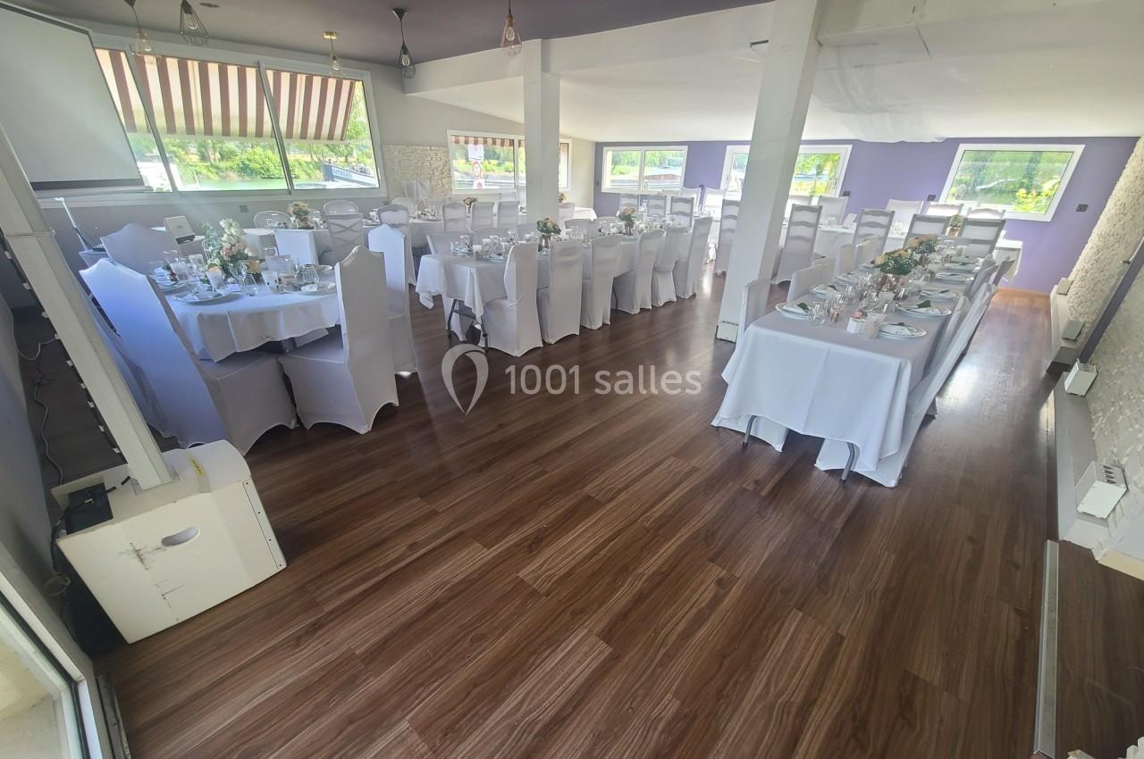 Salle de réception lumineuse avec tables dressées, nappes blanches et décorations florales, parquet en bois au sol.