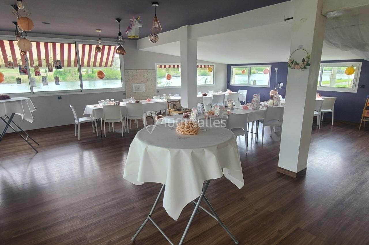 Salle lumineuse aménagée pour un événement, avec tables rondes dressées et décorations suspendues.