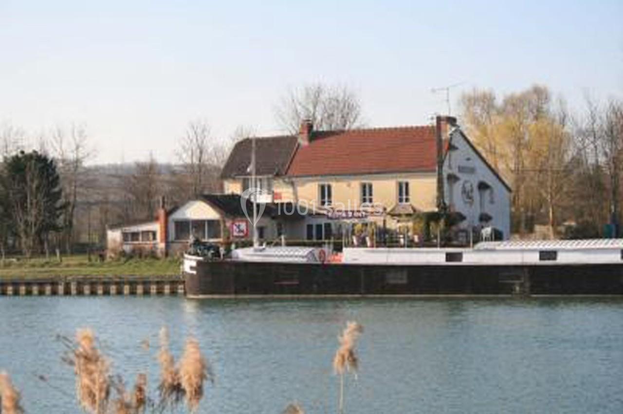Bâtiment avec terrasse au bord d'un canal, un bateau amarré au premier plan et des arbres en arrière-plan.