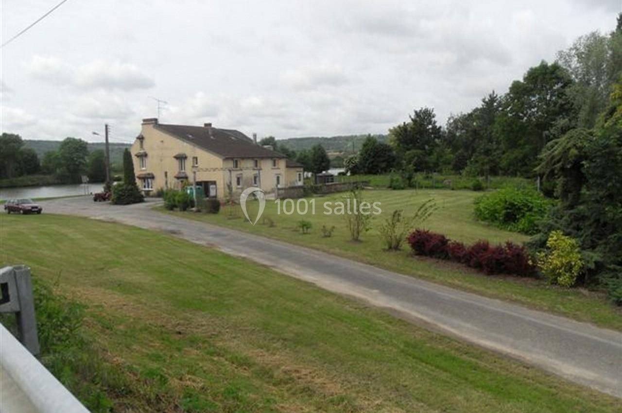 Maison en pierre avec jardin, située près d'une route et d'un plan d'eau, entourée de végétation et d'arbres.