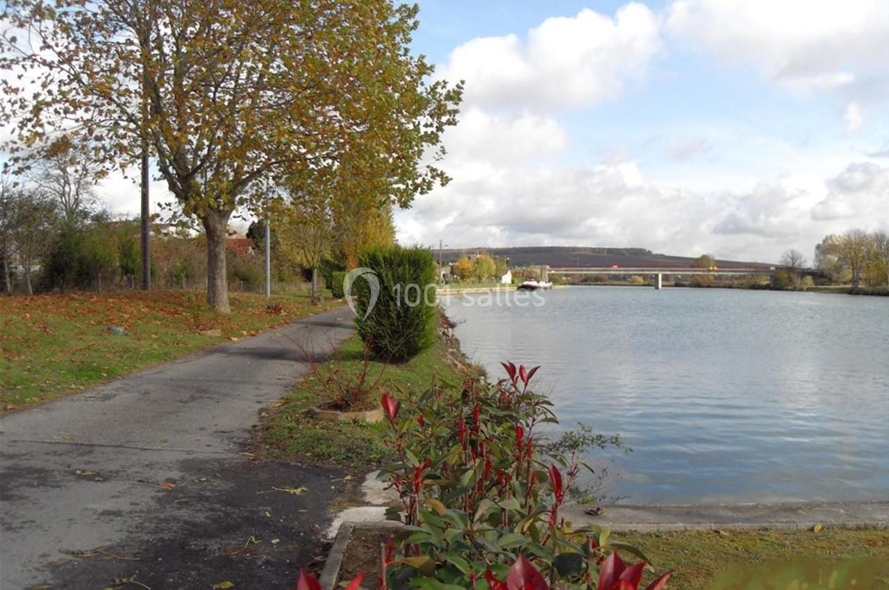 Chemin bordé d'arbres et de fleurs longeant une rivière sous un ciel partiellement nuageux.