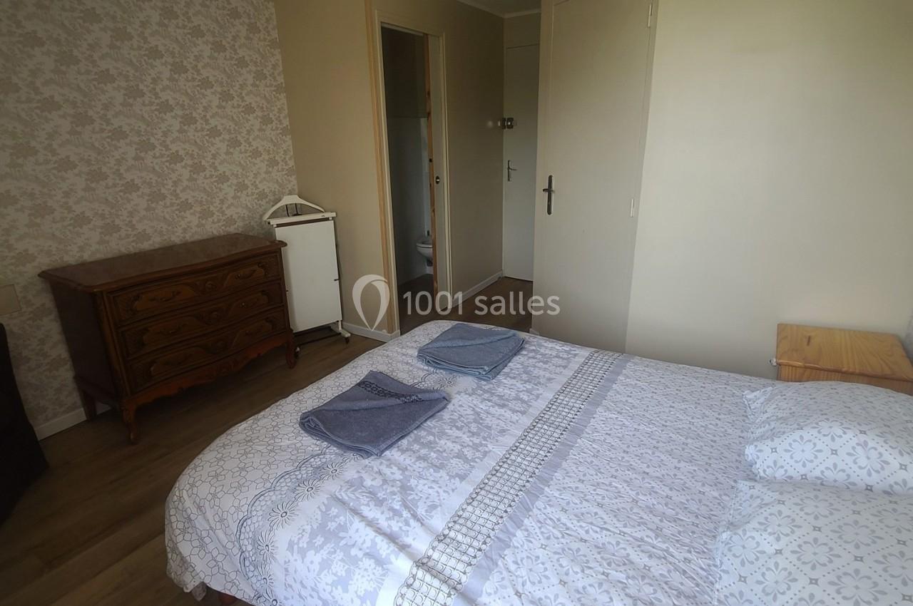 Chambre avec lit double, commode en bois, murs clairs et accès à une salle d'eau visible par une porte ouverte.