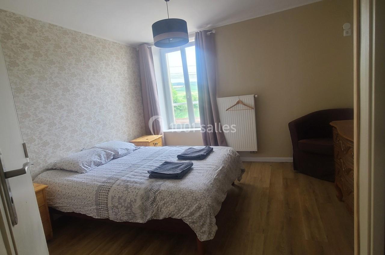 Chambre lumineuse avec lit double, tables de chevet, fauteuil, commode, fenêtre donnant sur un paysage verdoyant.