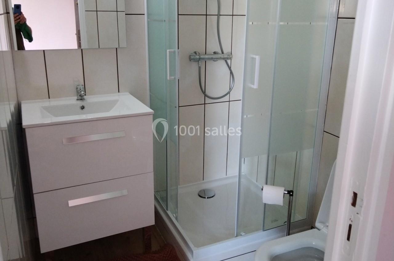 Salle de bain avec douche vitrée, meuble lavabo blanc, miroir et carrelage mural beige.