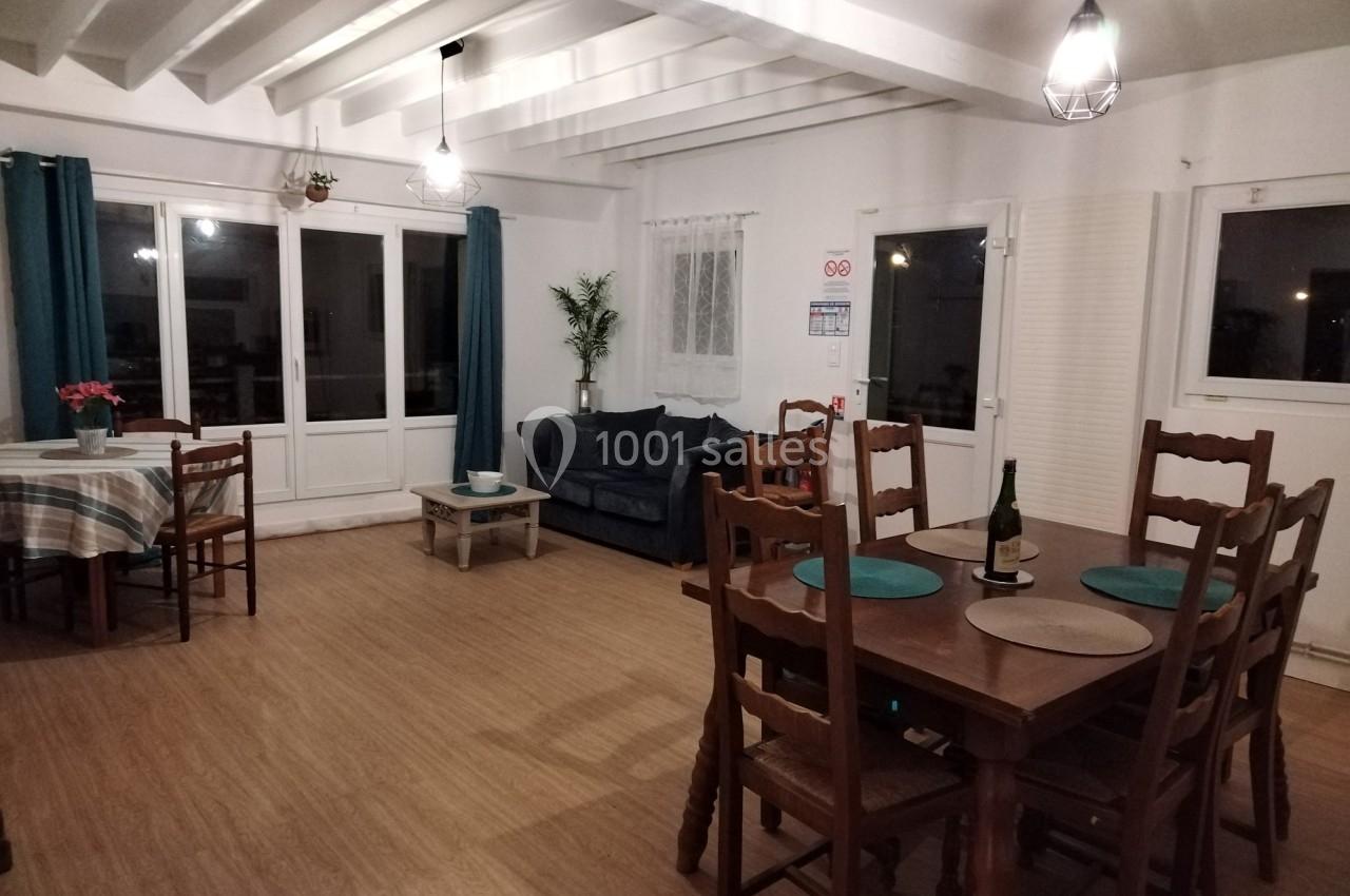 Salle à manger lumineuse avec table en bois, chaises, canapé, grandes fenêtres et éclairage suspendu.