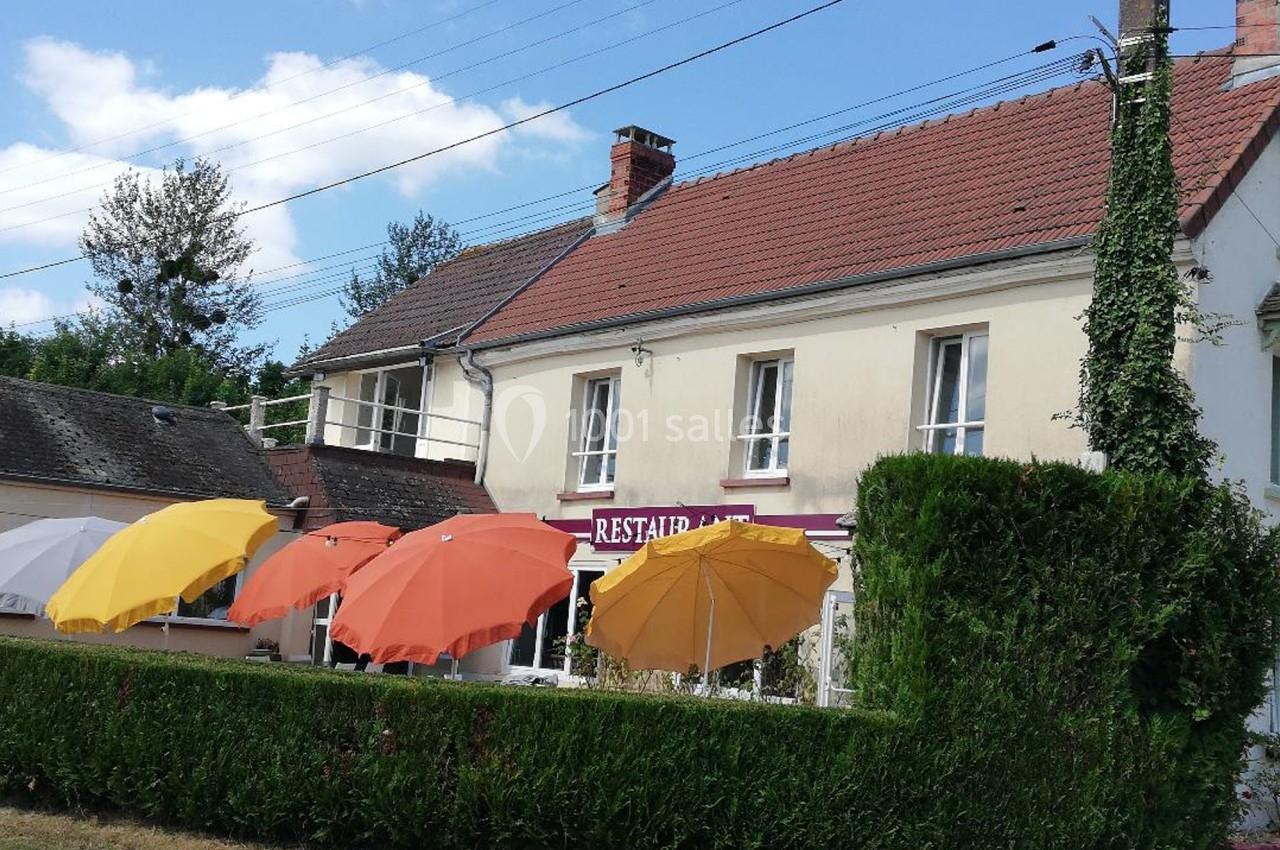 Façade d'un restaurant avec des parasols colorés en terrasse, entourée de haies et sous un ciel dégagé.