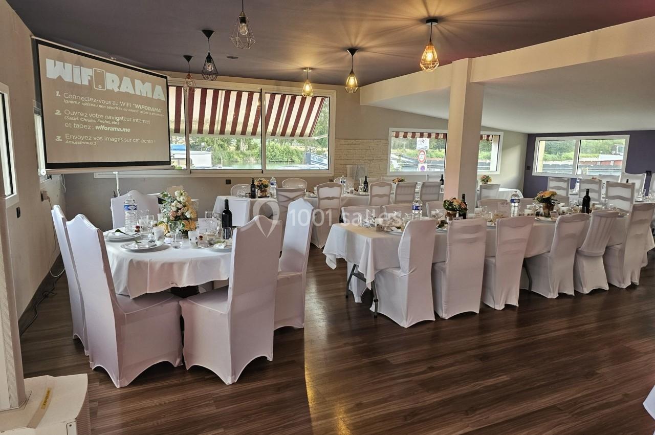 Salle de réception décorée pour un événement, avec tables rondes dressées et un écran de projection allumé.