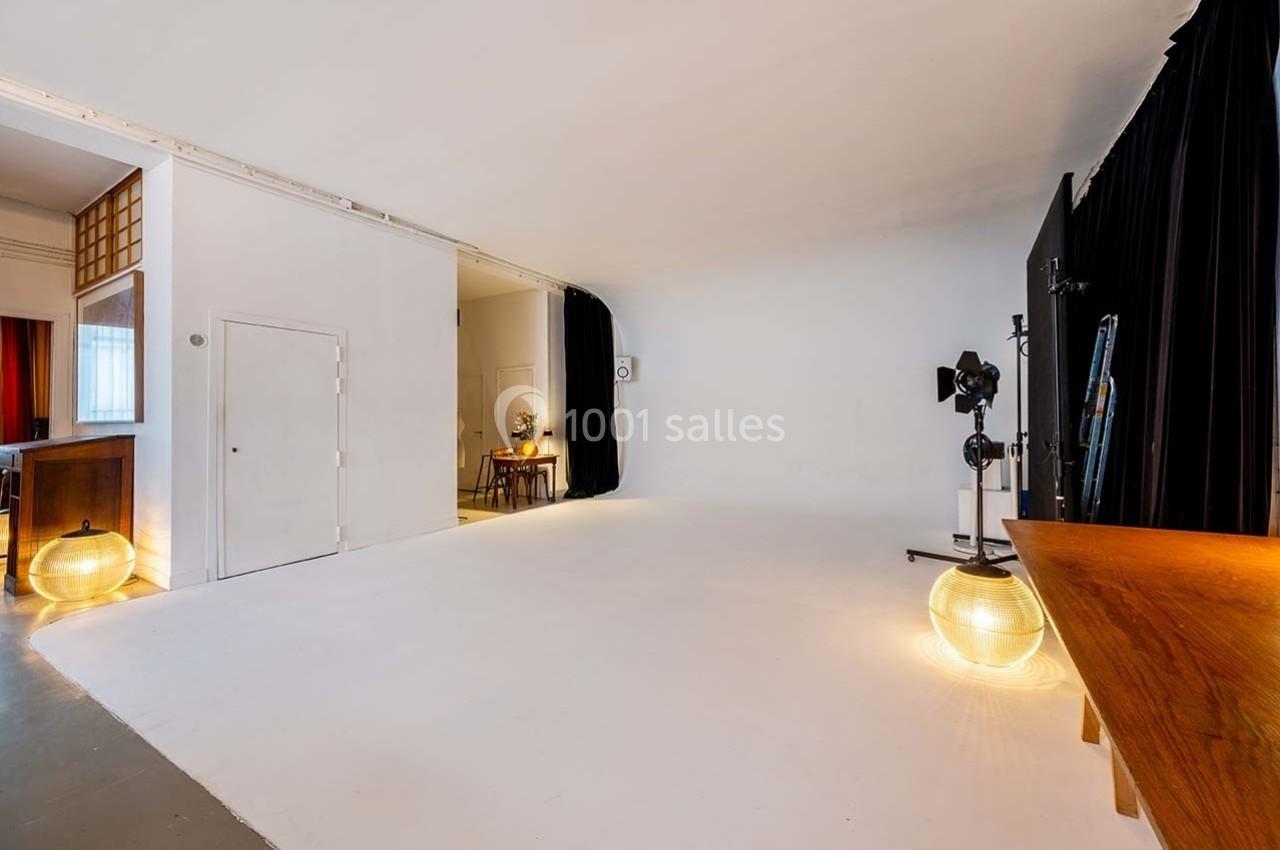 Studio lumineux avec fond blanc, éclairages, rideaux noirs et espace aménagé pour des prises de vue ou des tournages.