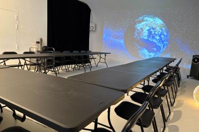 Salle avec des tables et chaises noires disposées en U, projection d’un paon coloré sur un mur blanc, rideaux noirs à droite.